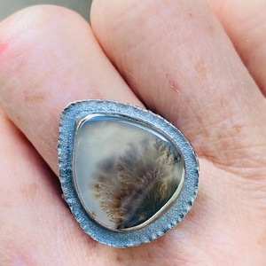 May include: A teardrop-shaped dendritic agate ring featuring a translucent stone with dark, fern-like inclusions. The gemstone is set within a textured, dark oxidized silver bezel with a rough, uneven edge that frames the central stone.