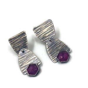 May include: A pair of silver earrings with a textured, striped design. Each earring features a geometric shape with a hexagonal, deep pink gemstone. The earrings are connected with small silver rivets.