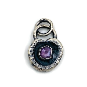 Puede incluir: Un colgante de plata con un marco ovalado texturizado. El colgante presenta un interior azul oscuro con una gema púrpura facetada en el centro. Dos bucles entrelazados forman la fianza en la parte superior.