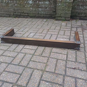 May include: A dark brown fireplace mantelpiece, possibly made of wood or metal, sits on a brick paved surface. The mantelpiece has a rectangular shape with decorative corner details. It appears to be an antique or reclaimed item, positioned outdoors.