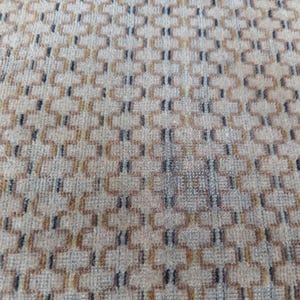 May include: Close-up of a beige rug with a repeating geometric pattern. The pattern features interconnected shapes in shades of brown and black, creating a textured surface. The rug appears to be made of a soft, woven material.