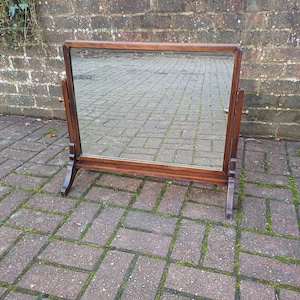 May include: A rectangular, wooden framed vanity mirror with a dark brown finish. The mirror is supported by two angled legs and a horizontal base. The mirror is standing on a brick surface.