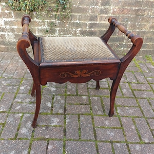 May include: A vintage wooden stool with a patterned seat and curved legs. The stool features decorative carvings on the apron and curved armrests. The wood has a dark finish, and the seat is upholstered.