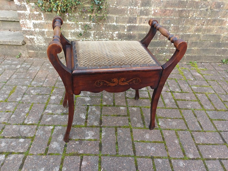 May include: A vintage wooden stool with a patterned seat and curved legs. The stool features decorative carvings on the apron and curved armrests. The wood has a dark finish, and the seat is upholstered.