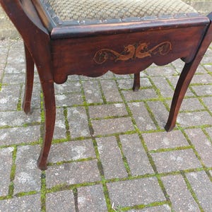 May include: A dark wooden chair with curved legs and a decorative apron. The chair has an ornate design on the apron and a patterned fabric seat. The chair is positioned on a brick surface. The chair's finish is a dark brown.