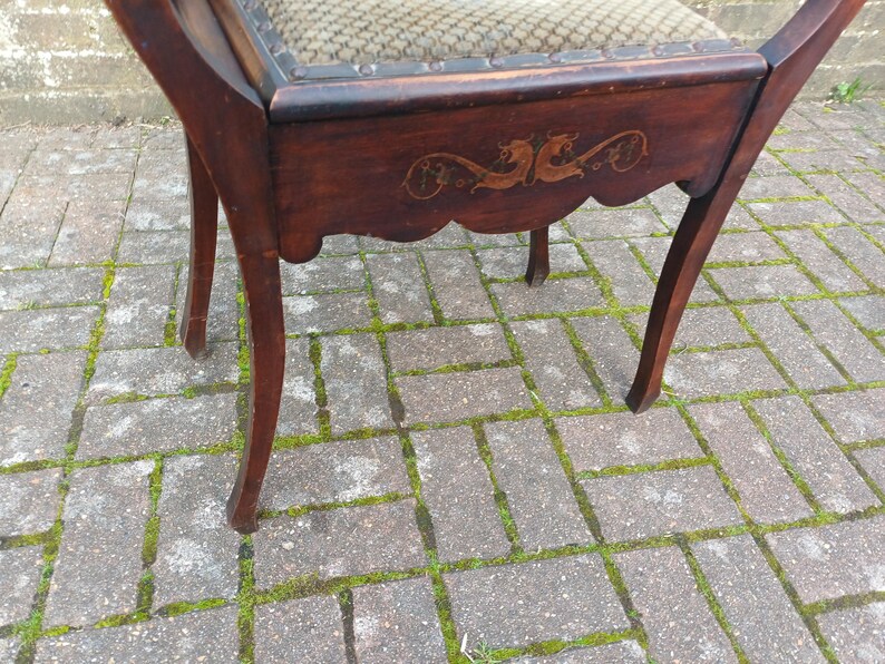 May include: A dark wooden chair with curved legs and a decorative apron. The chair has an ornate design on the apron and a patterned fabric seat. The chair is positioned on a brick surface. The chair's finish is a dark brown.