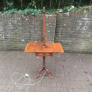 May include: An antique wooden lamp table with a small drawer and a tall lamp post. The table has a three-legged base and a rectangular top with drop-leaf sides. The table is made of warm-toned wood. The table's height is approximately 120 cm.