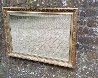 Op de afbeelding: Een gouden, sierlijke spiegel met een rechthoekige lijst hangt aan een bakstenen muur. De spiegel reflecteert een geplaveid pad.