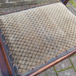 May include: Close-up of a vintage chair seat. The seat features a patterned fabric in tan and brown, framed by a dark wooden border. The border is secured with evenly spaced, round, metal studs. The chair's wooden frame is visible.