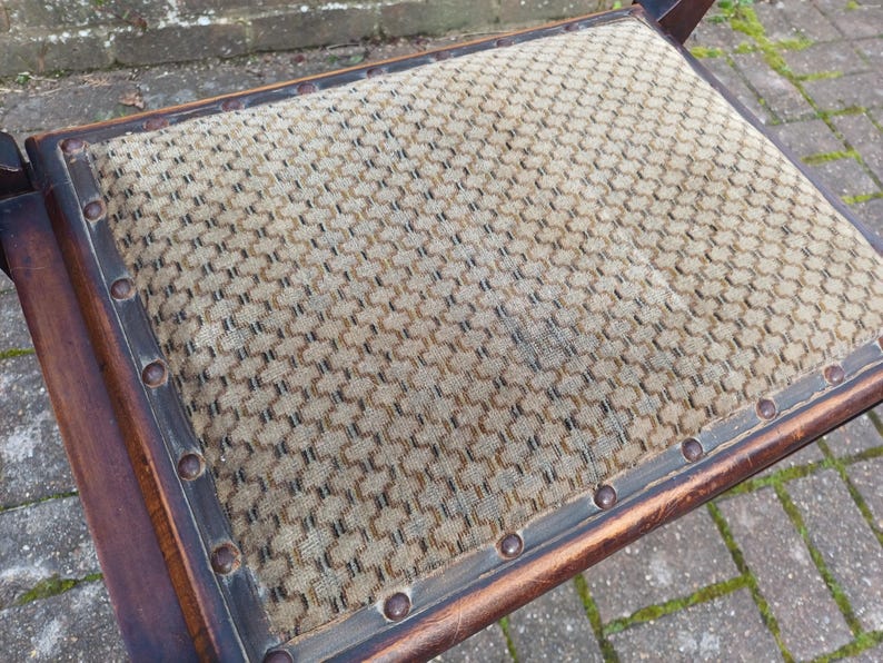 May include: Close-up of a vintage chair seat. The seat features a patterned fabric in tan and brown, framed by a dark wooden border. The border is secured with evenly spaced, round, metal studs. The chair's wooden frame is visible.