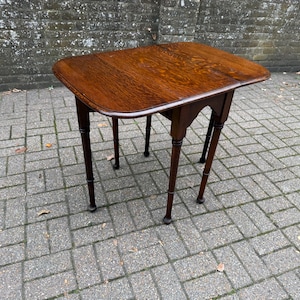 May include: A dark brown wooden drop-leaf table with a rectangular top and rounded corners. The table features four slender, turned legs with small, rounded feet and decorative arched supports connecting the legs to the tabletop frame.