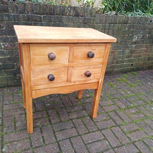 May include: A small, light-coloured wooden chest of drawers with four drawers and dark wooden knobs. The chest has a simple design and is supported by four legs. The wood has a natural finish.