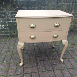 May include: A small, two-drawer chest in a neutral beige colour. The chest features curved drawers with brass-coloured handles and ornate, curved legs. The piece is likely a bedside table or small side table, adding a touch of vintage charm.