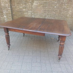 May include: A dark brown wooden table with a rectangular top and rounded corners. The table has four ornate, turned legs with small wheels. The table is an antique, with a classic design. The table is approximately 152 cm long and 91 cm wide.