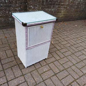 May include: White wicker cabinet with a glass top and a small door. The cabinet has a lavender trim and gold hardware. The cabinet is rectangular and stands on four small feet. The cabinet is outdoors on a brick surface.