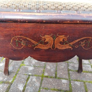 May include: Close-up of a vintage wooden chair with a dark brown finish. The chair features an ornate, decorative design in gold and green on the apron. The seat is upholstered with a patterned fabric. The chair's legs are slender and curved.