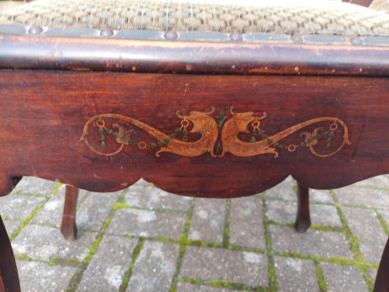 May include: Close-up of a vintage wooden chair with a dark brown finish. The chair features an ornate, decorative design in gold and green on the apron. The seat is upholstered with a patterned fabric. The chair's legs are slender and curved.