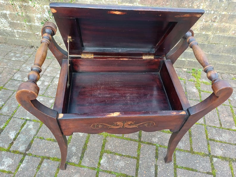 May include: An antique dark brown wooden side table with a hinged lid, revealing an open storage compartment. The table has curved legs and decorative armrests. The wood shows signs of age and wear.