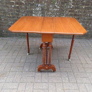 May include: A wooden drop-leaf table with a rectangular top and rounded corners. The table is a warm brown colour and has four legs with small wheels. The table is approximately 91 cm long when fully extended.
