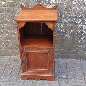 May include: A small, dark wood cabinet with a single door and a shelf. The cabinet has a decorative top with a carved design.
