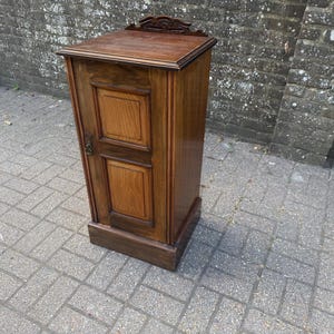 May include: A tall, rectangular wooden cabinet featuring a dark brown finish with lighter wood paneling on the single door. The cabinet has a flat top surface, a decorative carved wooden crest at the back, and a small metal handle on the left side.