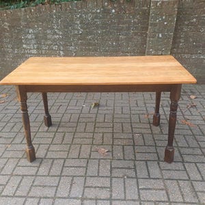 May include: A rectangular wooden table with a light brown tabletop and dark brown, turned legs. The table is set on a brick patio. The table is approximately 122 cm long and 76 cm wide. A brick wall is in the background.