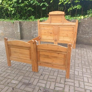 May include: A wooden bed frame, featuring a headboard and footboard. The headboard has a decorative top and paneling, while the footboard has a curved top. The bed frame is constructed from light-coloured wood.