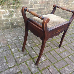 May include: A dark wood vanity stool with a patterned seat and curved legs. The stool has decorative carvings on the apron and a handle on each side. The seat is upholstered in a beige and brown fabric.