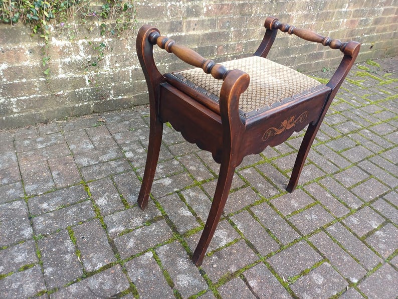 May include: A dark wood vanity stool with a patterned seat and curved legs. The stool has decorative carvings on the apron and a handle on each side. The seat is upholstered in a beige and brown fabric.