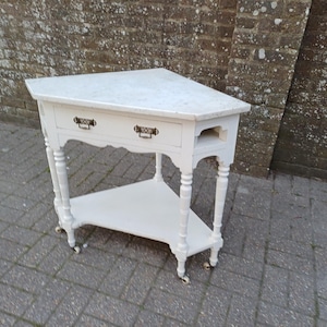 May include: A white corner table with a marble top, two drawers, and a lower shelf. The table has ornate legs and small wheels. The drawer handles are dark bronze. The table is painted white.
