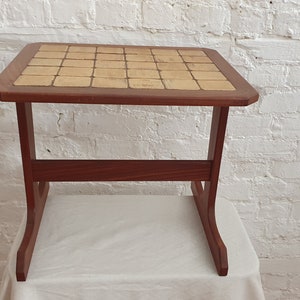 May include: A small, square wooden side table with a tiled top. The table has a dark brown frame and legs, and the tabletop is composed of beige tiles. The table is approximately 61 cm tall.
