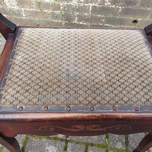 May include: An antique wooden chair with a woven seat. The chair has a dark brown finish with decorative carvings on the apron. The seat is a light tan colour with a geometric pattern and is framed by a dark border with metal studs.