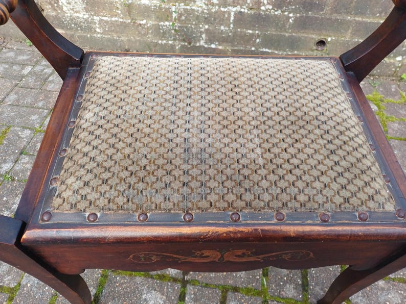 May include: An antique wooden chair with a woven seat. The chair has a dark brown finish with decorative carvings on the apron. The seat is a light tan colour with a geometric pattern and is framed by a dark border with metal studs.