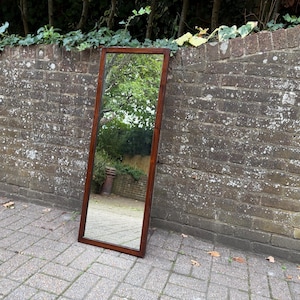 May include: A tall, rectangular floor mirror featuring a simple, dark brown wooden frame. The mirror surface is clear and reflective, showing a view of trees and foliage. The frame has straight, clean edges and a smooth, polished finish.