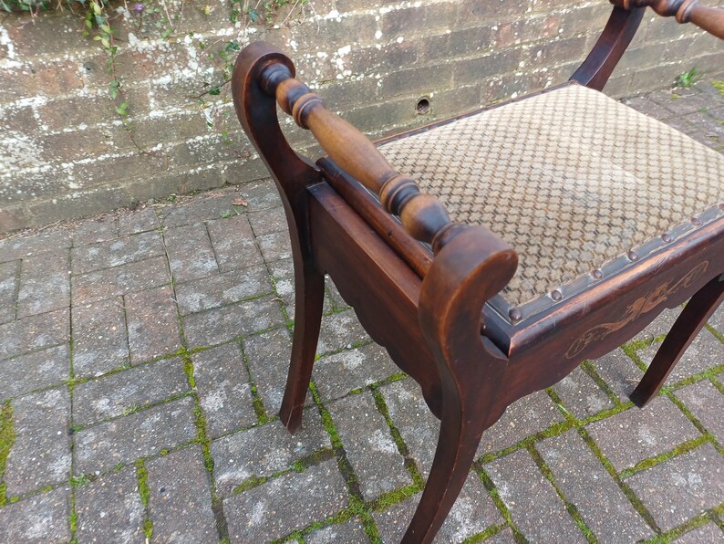 May include: An antique wooden chair with a patterned seat and curved armrests. The chair features dark brown wood with decorative carvings and a light-coloured patterned fabric. The legs are curved, and the chair appears well-preserved.