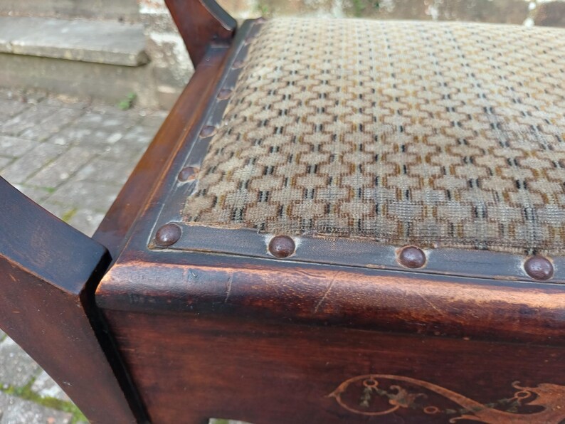 May include: Close-up of a vintage wooden chair with a patterned seat. The dark brown wood frame features decorative carvings and metal studs along the seat's edge. The seat has a beige and brown geometric pattern.