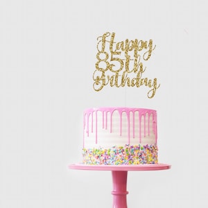 May include: Gold glitter cake topper with the text "Happy 85th Birthday" on a pink and white cake with sprinkles.