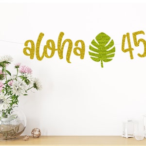 May include: Gold glitter banner with the words "Aloha" and the number "45" with a green glitter tropical leaf in between.