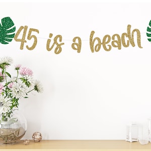 May include: Gold glitter banner with the text "45 is a beach" and green glitter tropical leaves on either end.