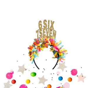 May include: A festive headband with colorful paper fringe and a gold glitter topper that reads "6 SIX 7 SEVEN". The headband is black and surrounded by confetti and star-shaped decorations in various colors.