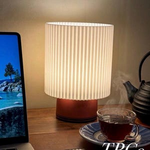 May include: A modern table lamp with a pleated white shade and a red base. The lamp is on a wooden desk next to a laptop, a cup of tea, and a teapot. The text "TBCo." is visible.
