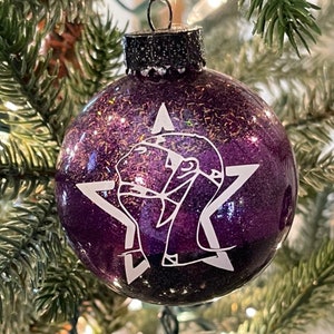 May include: A spherical purple Christmas ornament featuring a white graphic design of a star with a stylized figure inside. The ornament has a glittery finish and a black cap at the top with a small metal loop for hanging on a tree branch.