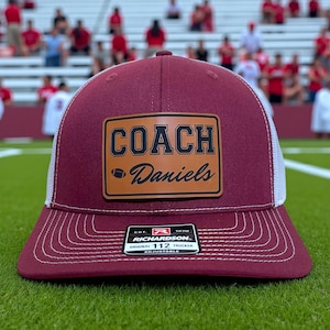 May include: Maroon and white trucker hat with a leather patch that reads "COACH Daniels" with a football graphic. The hat has a curved brim and white mesh back. The hat is adjustable.