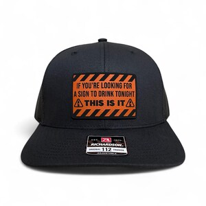 May include: Black trucker hat with a black mesh back and a curved brim. The hat features a rectangular patch with the text "IF YOU'RE LOOKING FOR A SIGN TO DRINK TONIGHT THIS IS IT" in orange and white. The hat is adjustable.