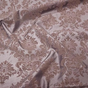 May include: Close-up of a luxurious fabric with a damask pattern. The fabric is a soft lavender color with a repeating design in a muted brown tone. The fabric has a subtle sheen and is draped, showing folds and texture.