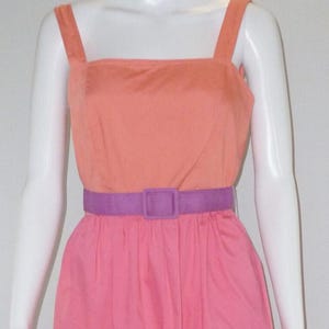 May include: A two-tone dress in peach and pink, featuring a square neckline and wide straps. A purple belt cinches the waist. The dress is knee-length and made of a smooth fabric.