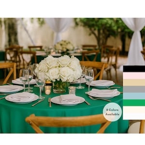 May include: A formal table setting with a vibrant emerald green tablecloth. The table is set with white plates, napkins, silverware, and wine glasses. A centerpiece of white roses sits in a clear vase. Wooden chairs surround the table.