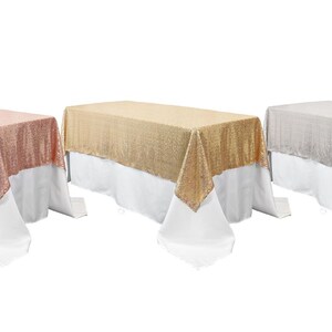May include: Three rectangular tablecloths in rose gold, gold, and silver. Each tablecloth is made of a shimmering sequined fabric and has a white lining.