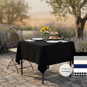 May include: A black square tablecloth covers a metal table set for dining outdoors. The table is set with white plates, silverware, and a vase of sunflowers. A wrought iron chair sits nearby. Six colors are available.
