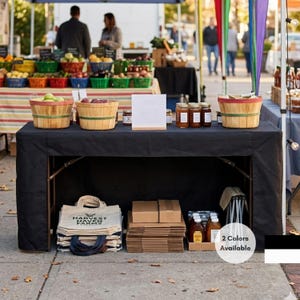May include: A black rectangular table cover draped over a folding table at an outdoor market. The table holds wooden baskets of produce, glass jars, and a sign. Underneath, there are stacks of canvas tote bags, cardboard boxes, and honey jars.